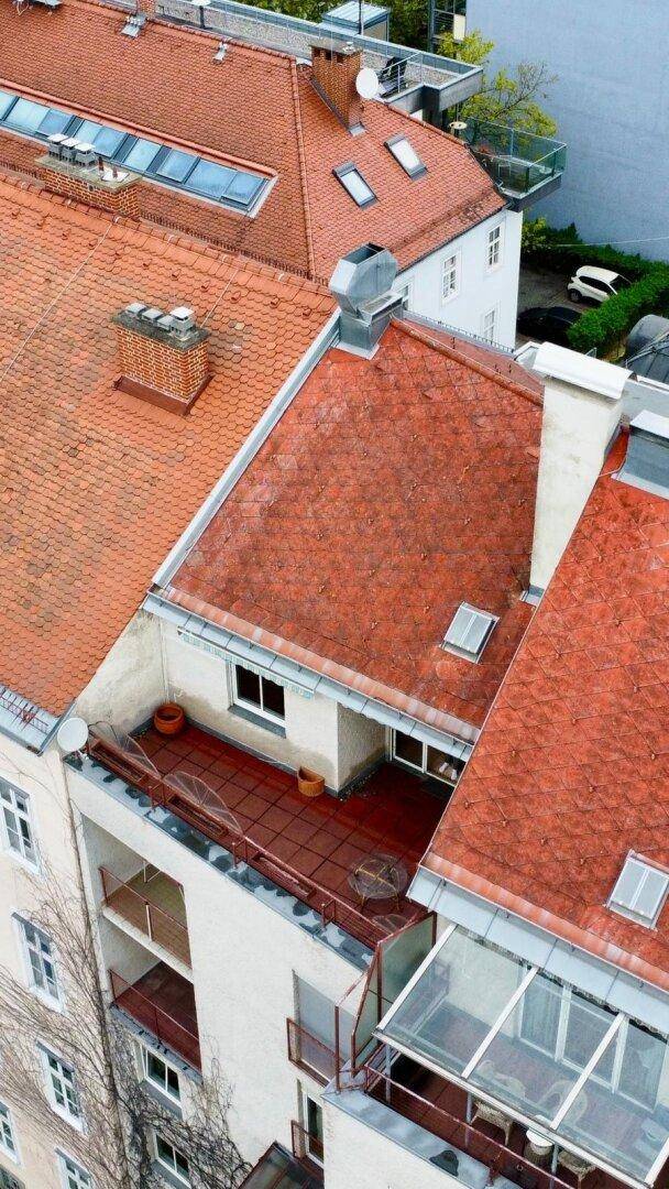 Dachlandschaft mit roten Ziegeldächern, Dachfenstern und einer Dachterrasse in urbaner Umgebung.