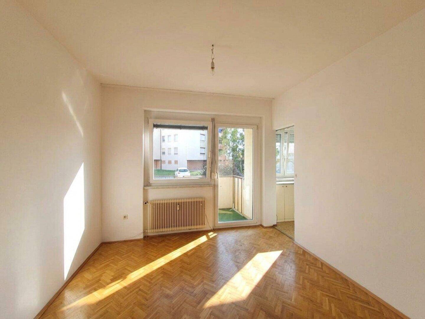 Lichtdurchflutetes Wohnzimmer mit Parkettboden und Zugang zu einem kleinen Balkon mit Kunstrasen.