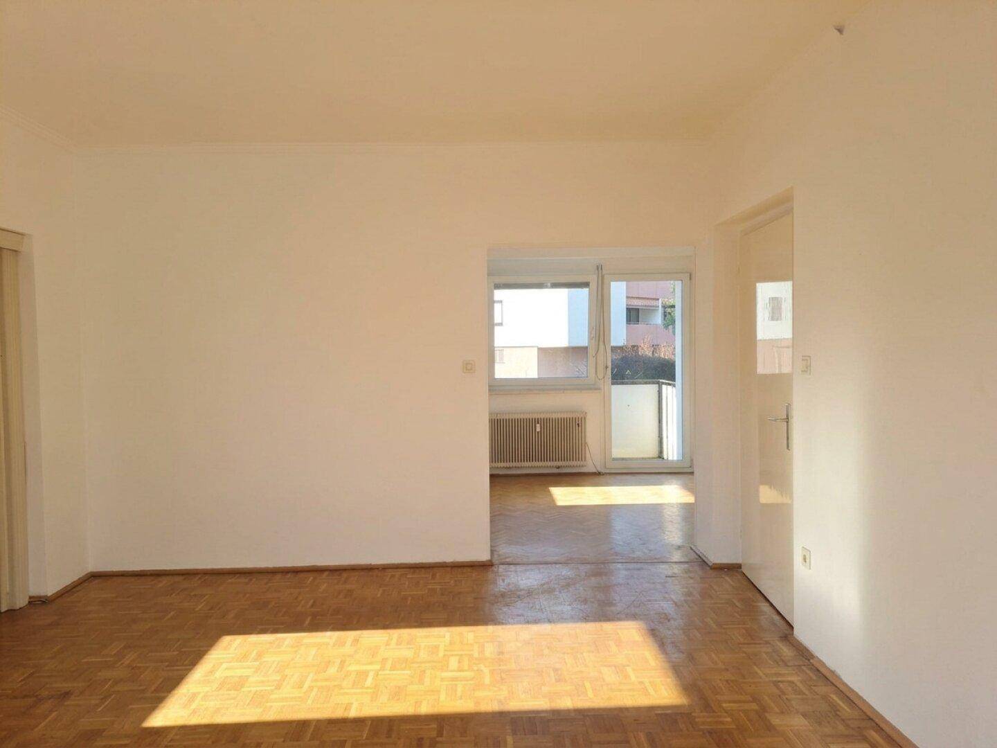 Geräumiges Wohnzimmer mit Parkettboden und viel Tageslicht, das in den angrenzenden Bereich führt.