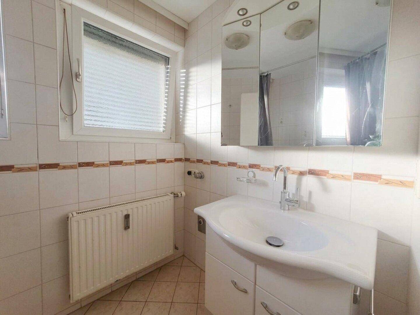 Badezimmer mit Fliesenboden, Waschbeckenunterschrank, Spiegelschrank und Fenster für Tageslicht.