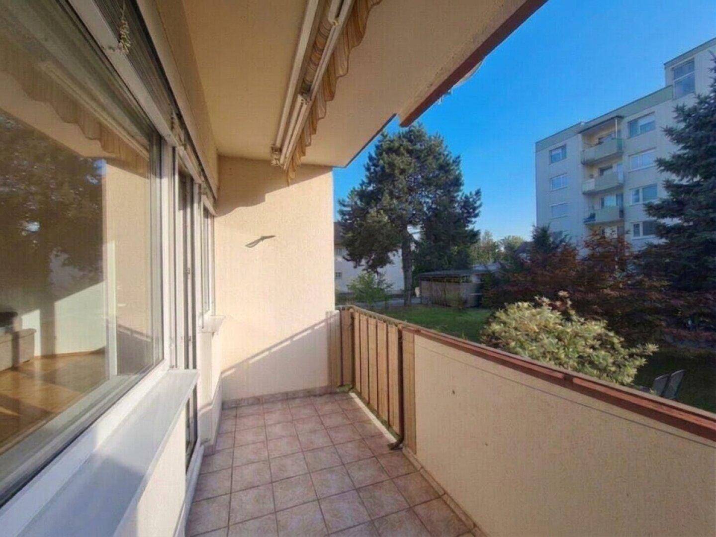 Heller Balkon mit Fliesenboden und Blick auf den grünen Innenhof und das Nachbargebäude.