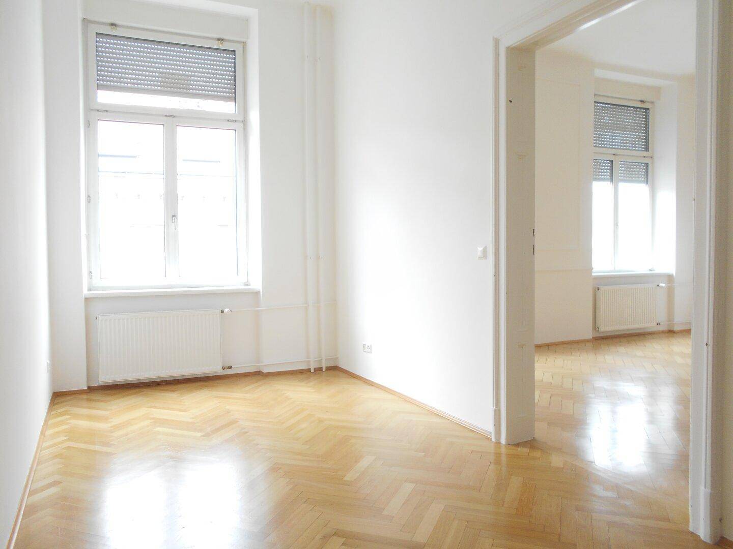 Heller Wohnbereich mit Fischgrätparkett und Blick durch eine offene Tür in einen weiteren Raum mit Fenstern.