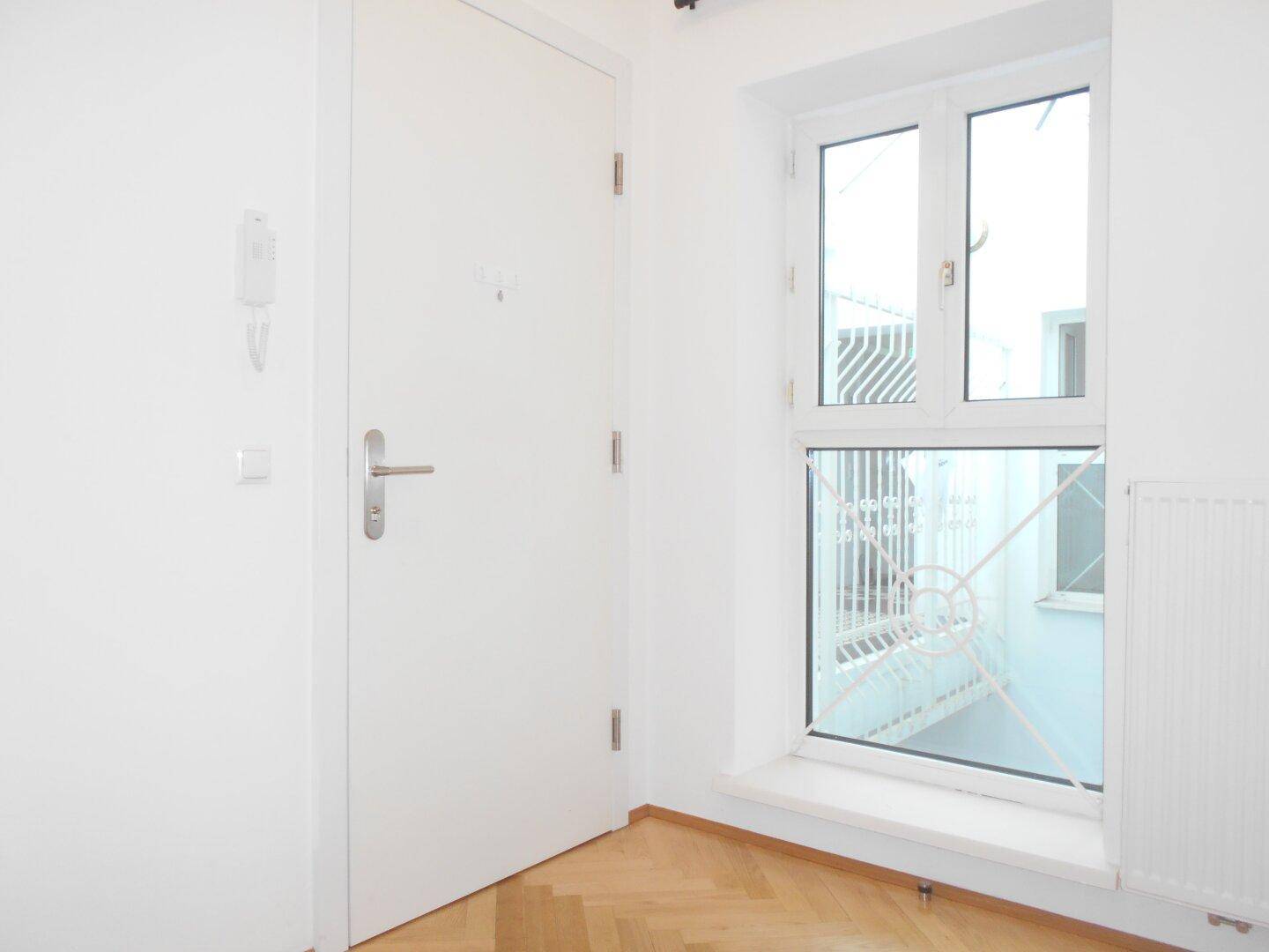 Eingangsbereich mit weißer Wohnungstür, Gegensprechanlage und einem Fenster mit Blick auf den Innenhof.