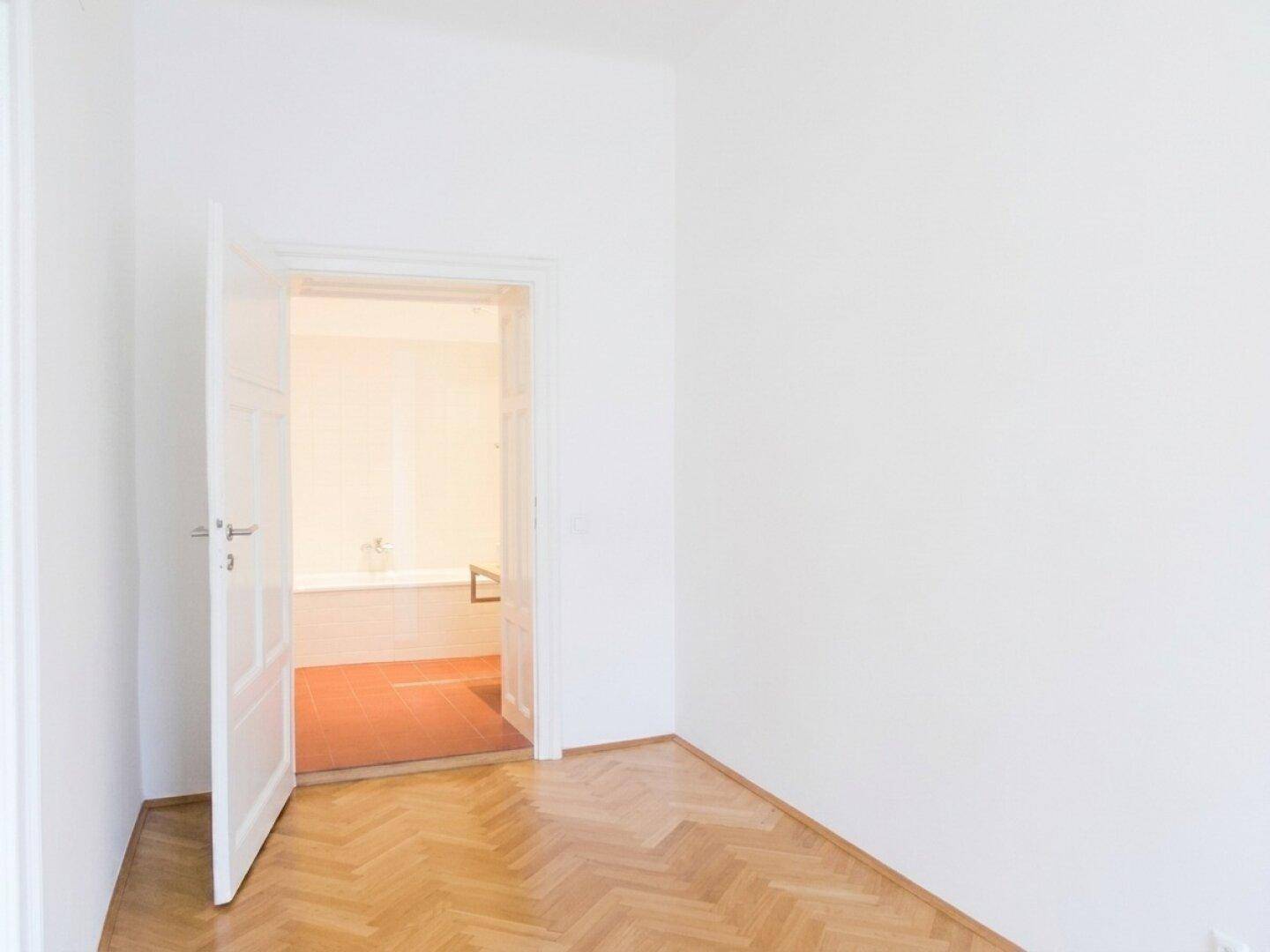 Heller Raum mit Fischgrätparkett und Blick durch eine offene Tür in ein Badezimmer mit Badewanne.