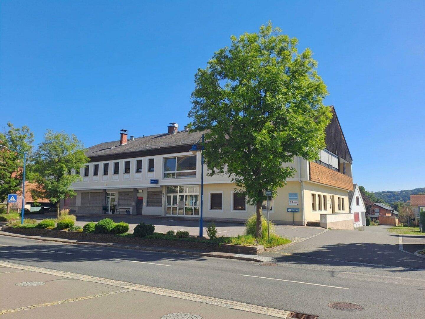 Gebäudeansicht mit Geschäften im Erdgeschoss und Wohnungen darüber, umgeben von Grünflächen.