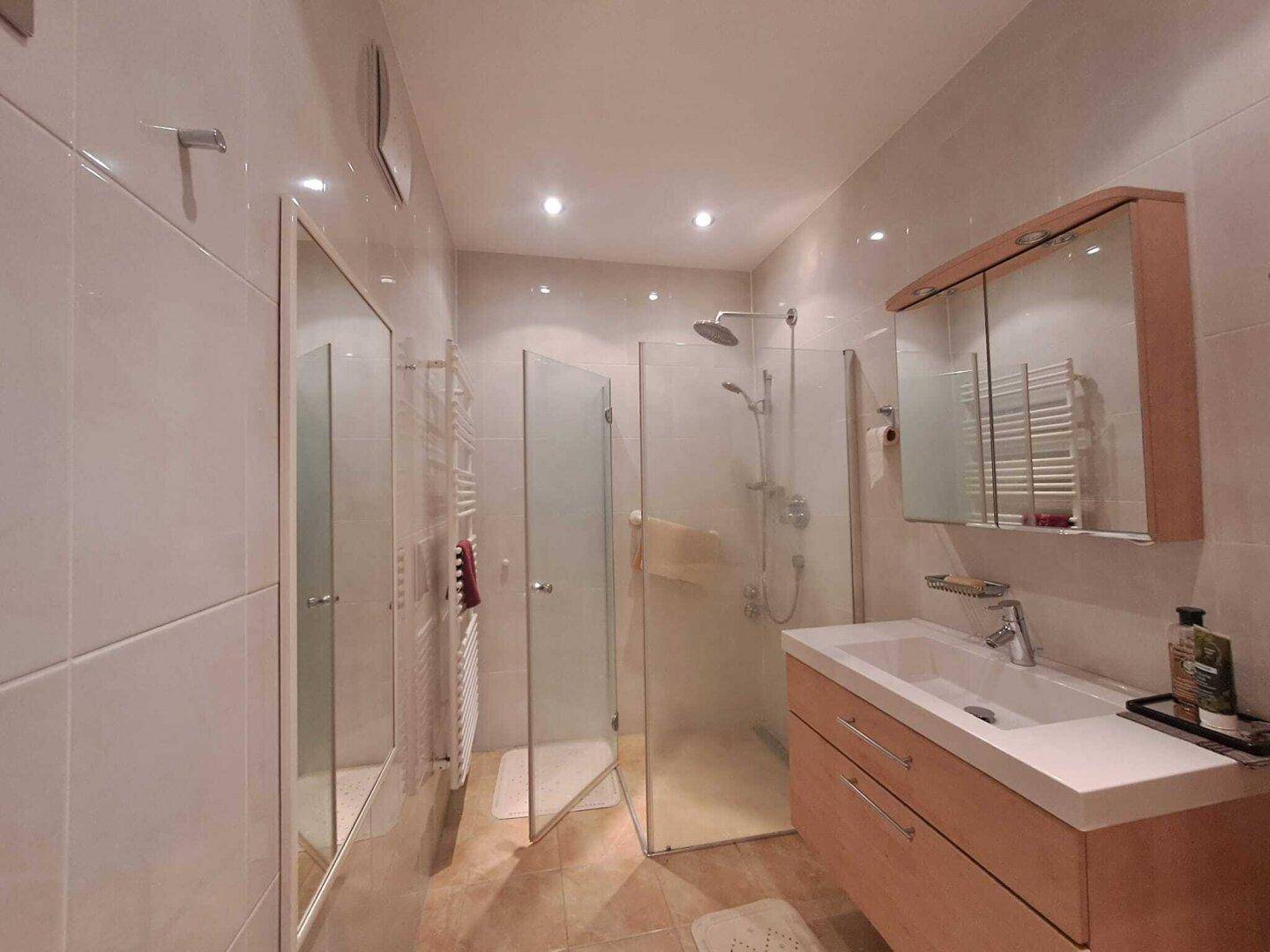 Helles Badezimmer mit moderner Dusche, Waschtisch und Spiegelschrank, gut beleuchtet.