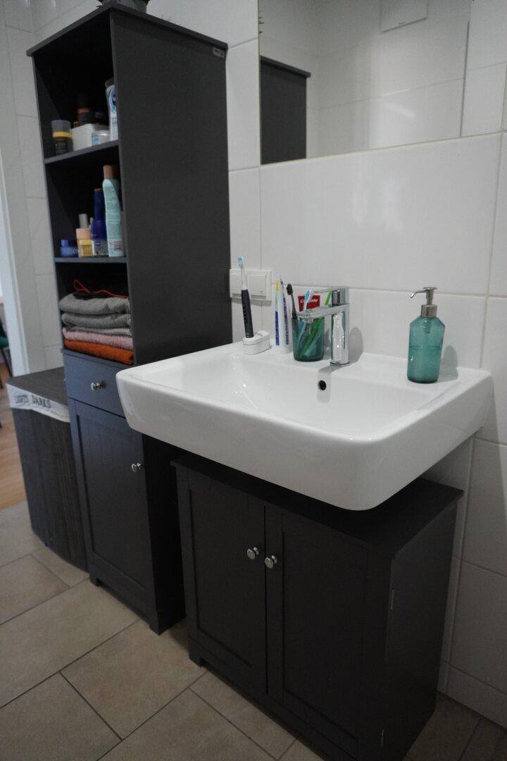 Modernes Badezimmer mit weißem Waschbecken, dunklen Schränken und weißen Fliesen.