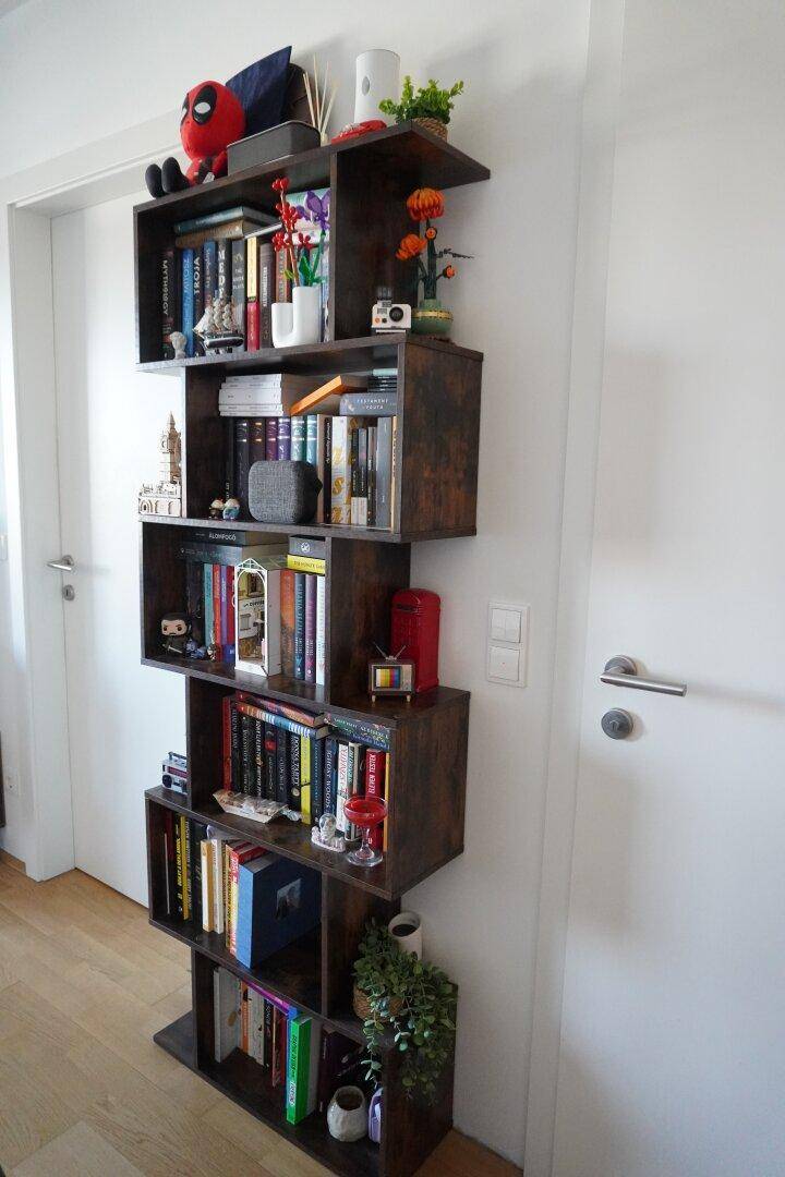 Hohes, schmales Bücherregal mit vielen Büchern und Dekorationen neben einer Tür.