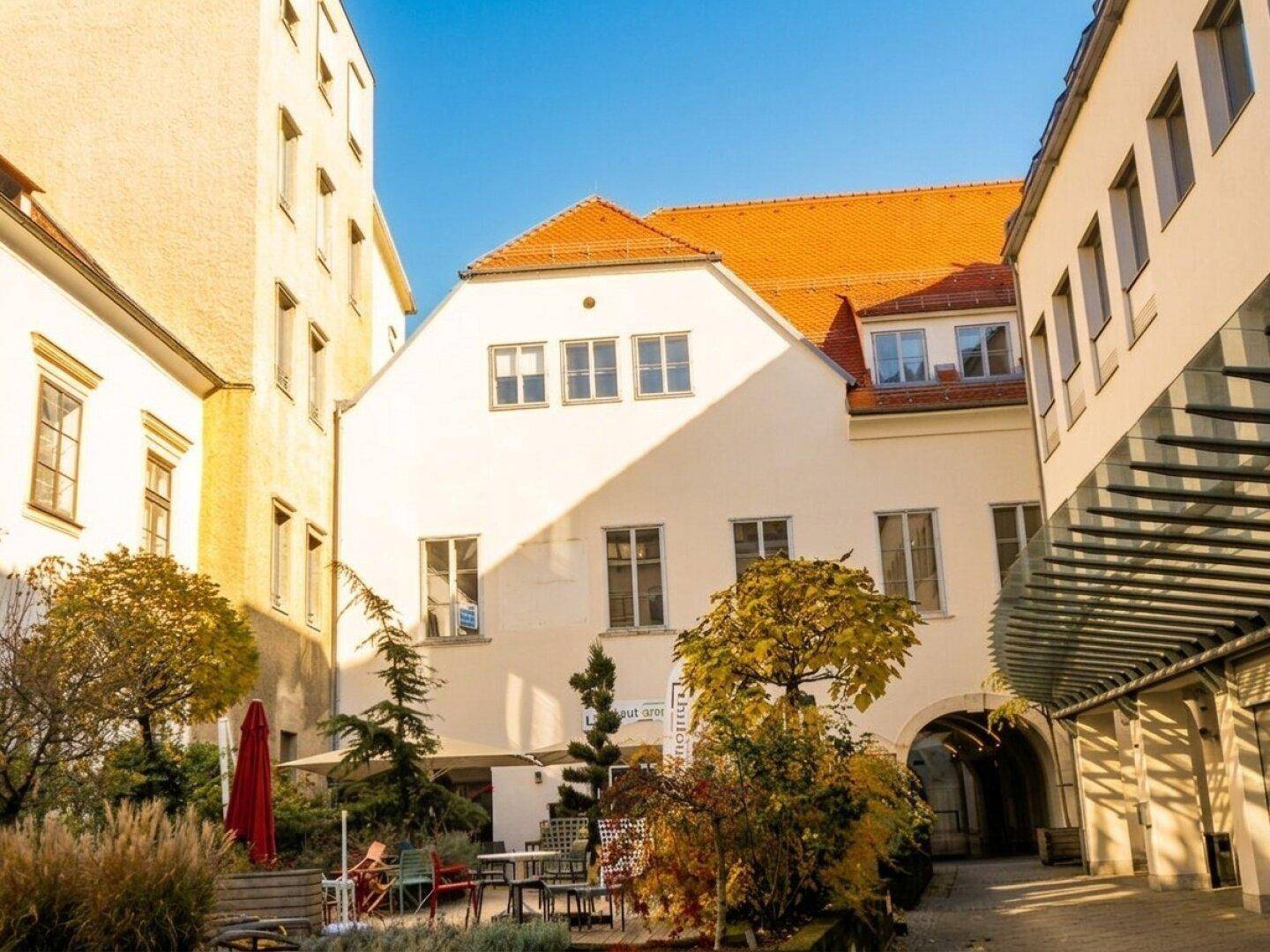 Malerischer Innenhof mit Sitzgelegenheiten, Bäumen und einem Gebäude mit rotem Ziegeldach im Hintergrund.