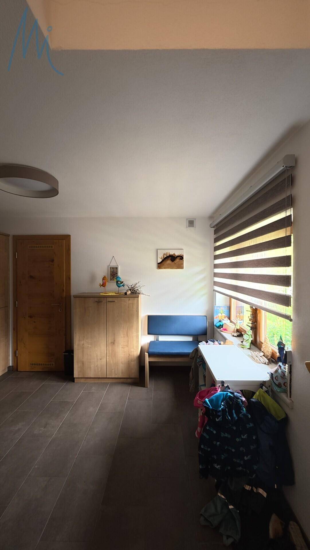 Heller Flur mit dunklem Fliesenboden, Fenster mit Jalousien und einer Sitzbank.