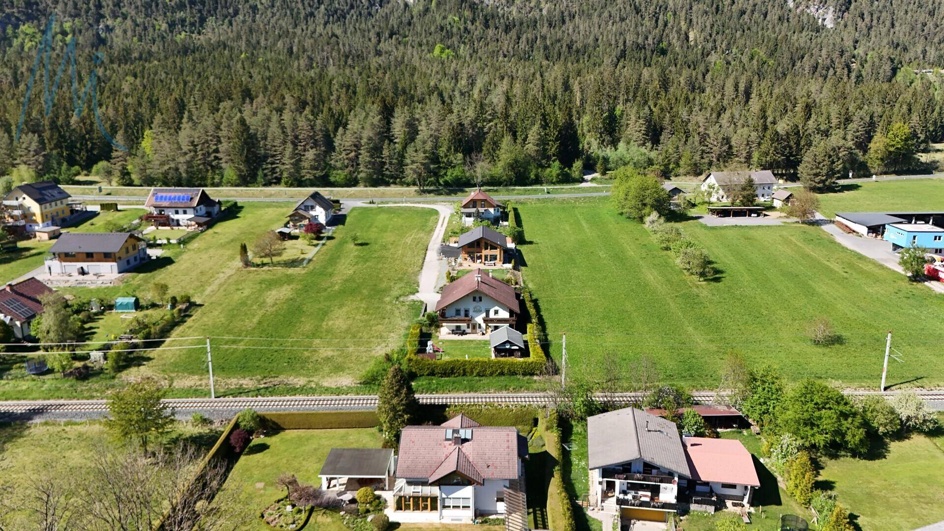 Weitläufige Luftaufnahme einer ländlichen Siedlung mit mehreren Häusern, grünen Feldern und Wald im Hintergrund.