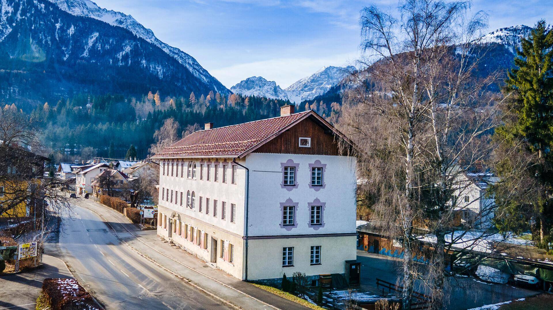 Großes Gebäude in alpiner Landschaft mit schneebedeckten Bergen im Hintergrund und einer Straße davor.