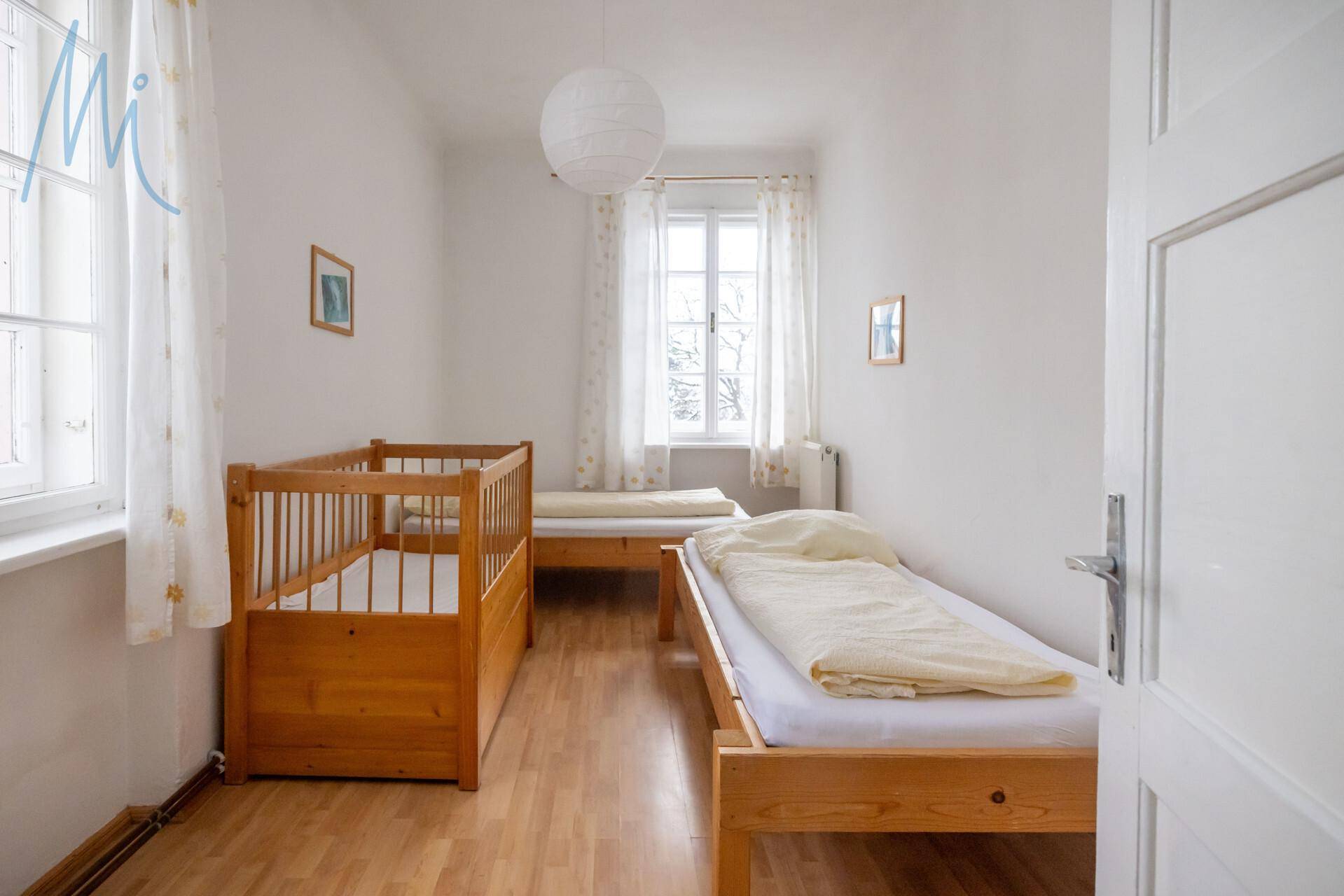 Helles Schlafzimmer mit einem Einzelbett und einem Babybett aus Holz, großen Fenstern und weißen Vorhängen.