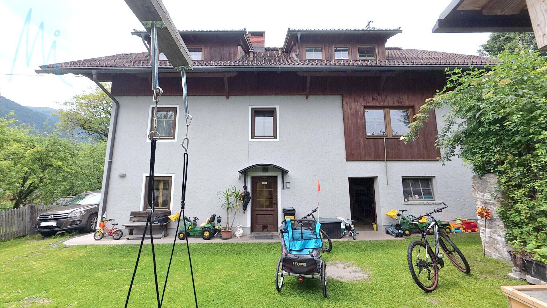 Vorderansicht des Hauses mit Eingangsbereich, Rasenfläche und Blick auf die umliegende Berglandschaft.