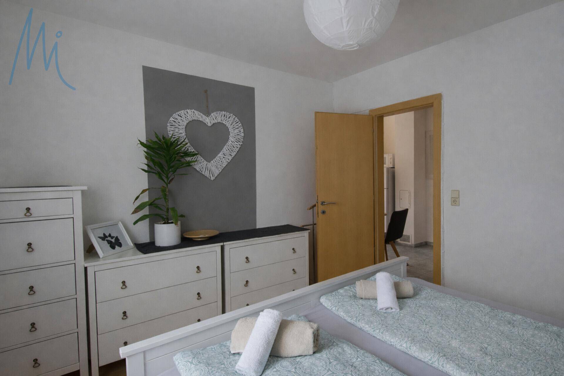 Helles Schlafzimmer mit Doppelbett, Kommoden und dekorativem Herz-Wandobjekt.