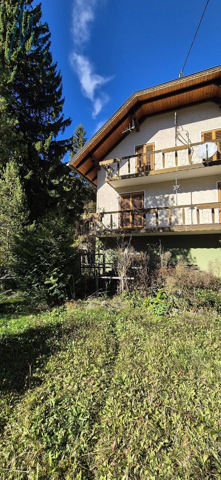 Seitenansicht eines Hauses mit Balkonen, umgeben von dichter Vegetation und Bäumen unter blauem Himmel.