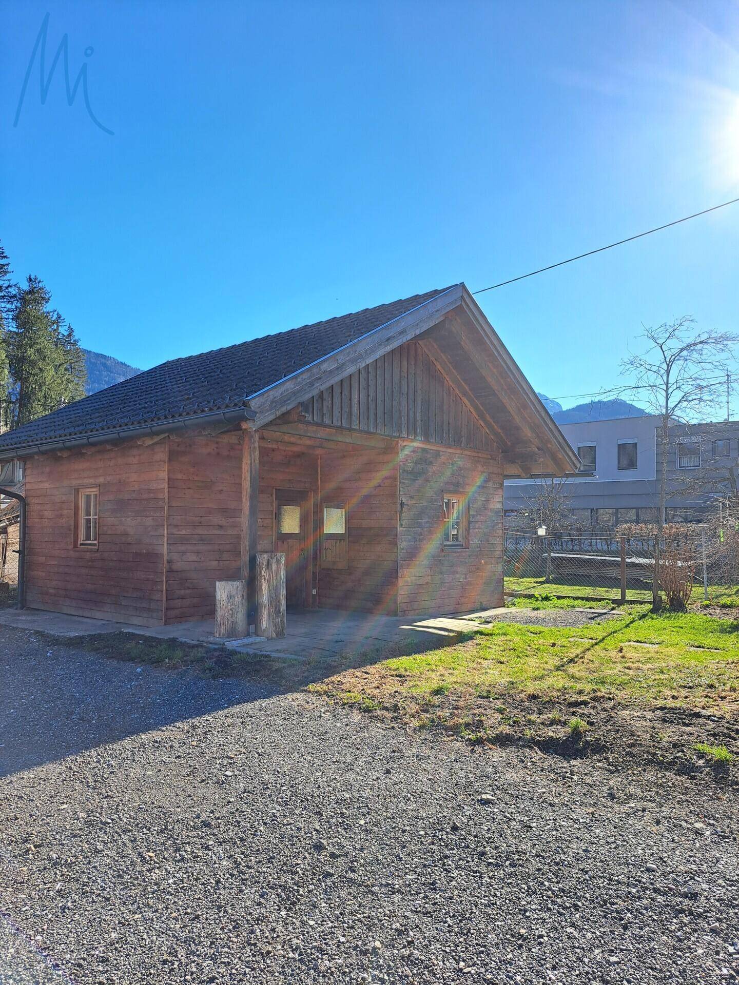 Rustikales Holzgebäude mit Vordach und angrenzender Grünfläche unter strahlendem Himmel.