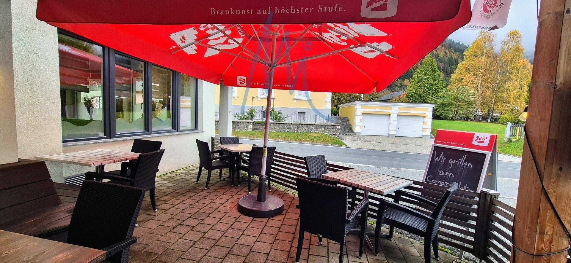 Überdachte Außenterrasse mit roten Sonnenschirmen, Holztischen und Stühlen, umgeben von Bäumen.