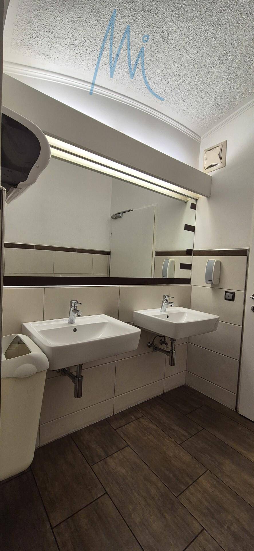 Waschbereich mit zwei modernen Waschbecken, großem Spiegel und Fliesenboden in einer öffentlichen Toilette.