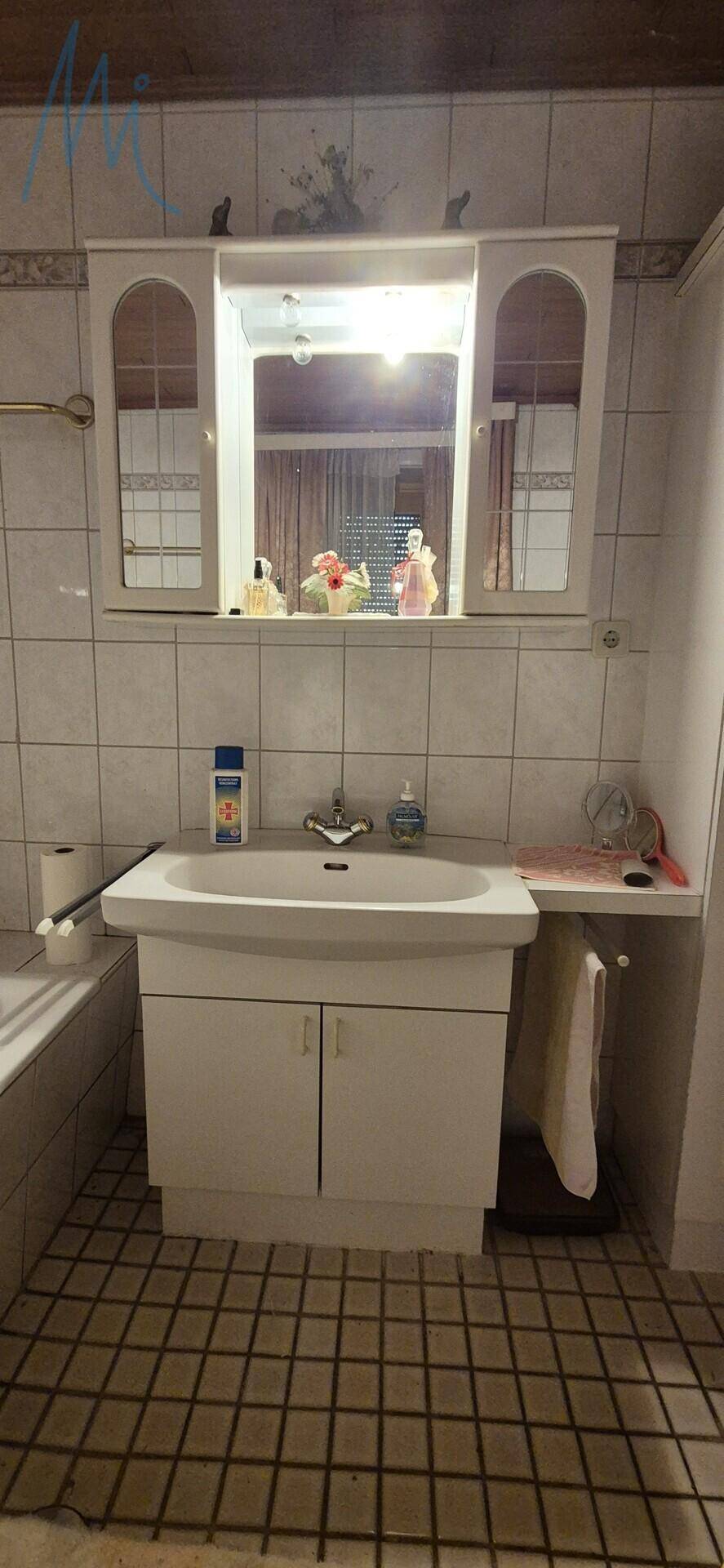 Badezimmer mit weißem Waschbecken, Spiegelschrank und hellen Fliesen an Wänden und Boden.