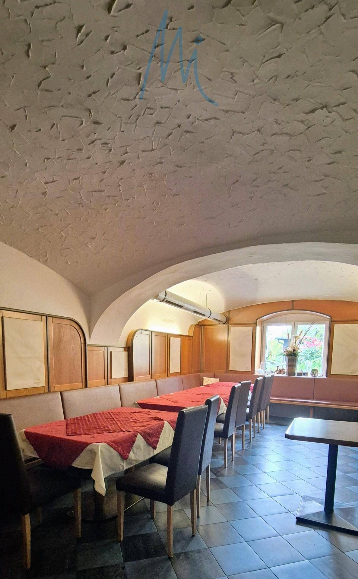 Langer Restaurantbereich mit gepolsterten Sitzbänken, roten Tischdecken und Fliesenboden unter einer strukturierten Decke.