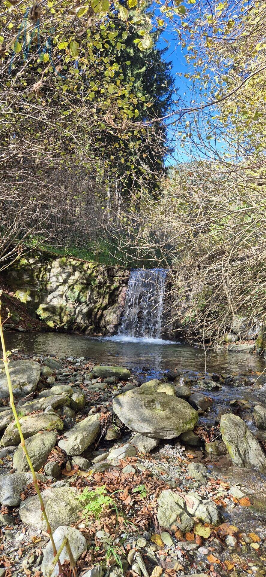 Kleiner Wasserfall in einer natürlichen Umgebung mit Felsen, Bäumen und klarem Wasser.