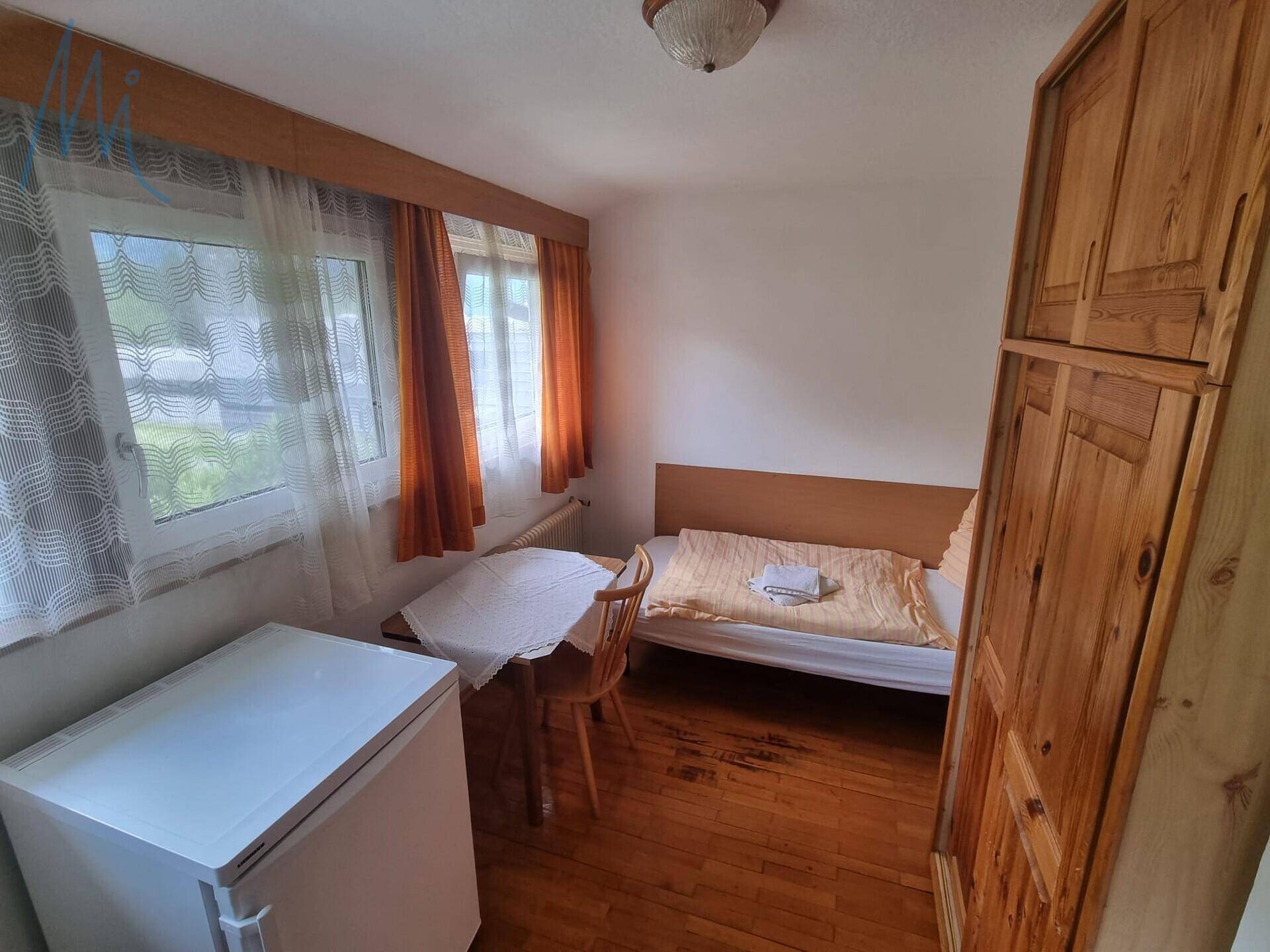 Helles Schlafzimmer mit Holzboden, Einzelbett, Tisch und einem Kühlschrank.