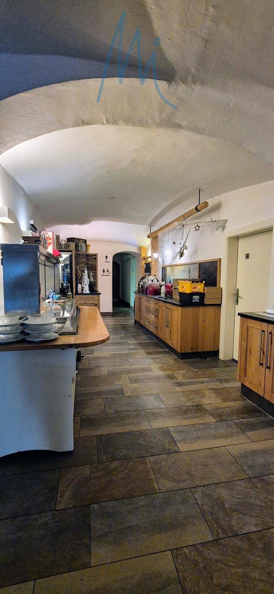 Langer Raum mit gewölbter Decke, Fliesenboden und Holzschränken, genutzt für Vorbereitung und Lagerung.