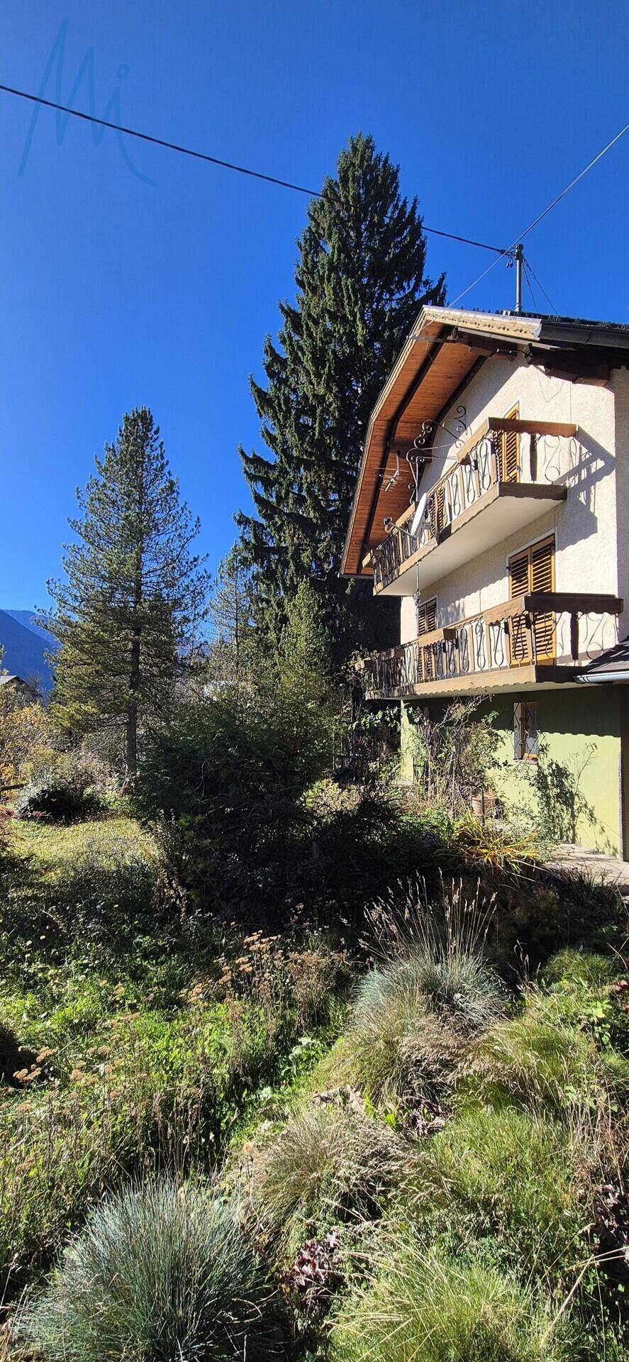 Seitenansicht des Hauses mit mehreren Holzbalkonen und üppiger Vegetation unter blauem Himmel.