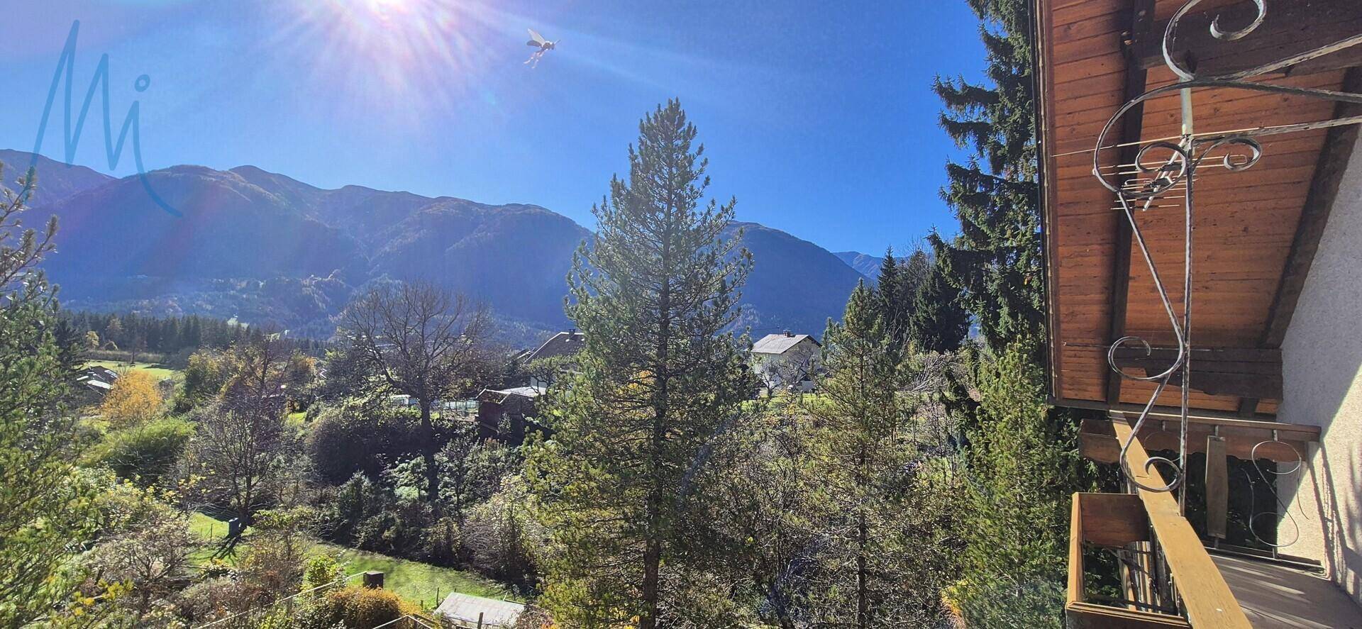 Panoramablick vom Balkon auf Berge, Bäume und Häuser unter strahlend blauem Himmel.