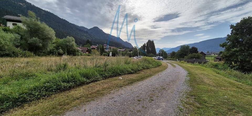 Schotterweg führt durch eine grüne Landschaft mit Häusern, Bäumen und Bergen in der Ferne.