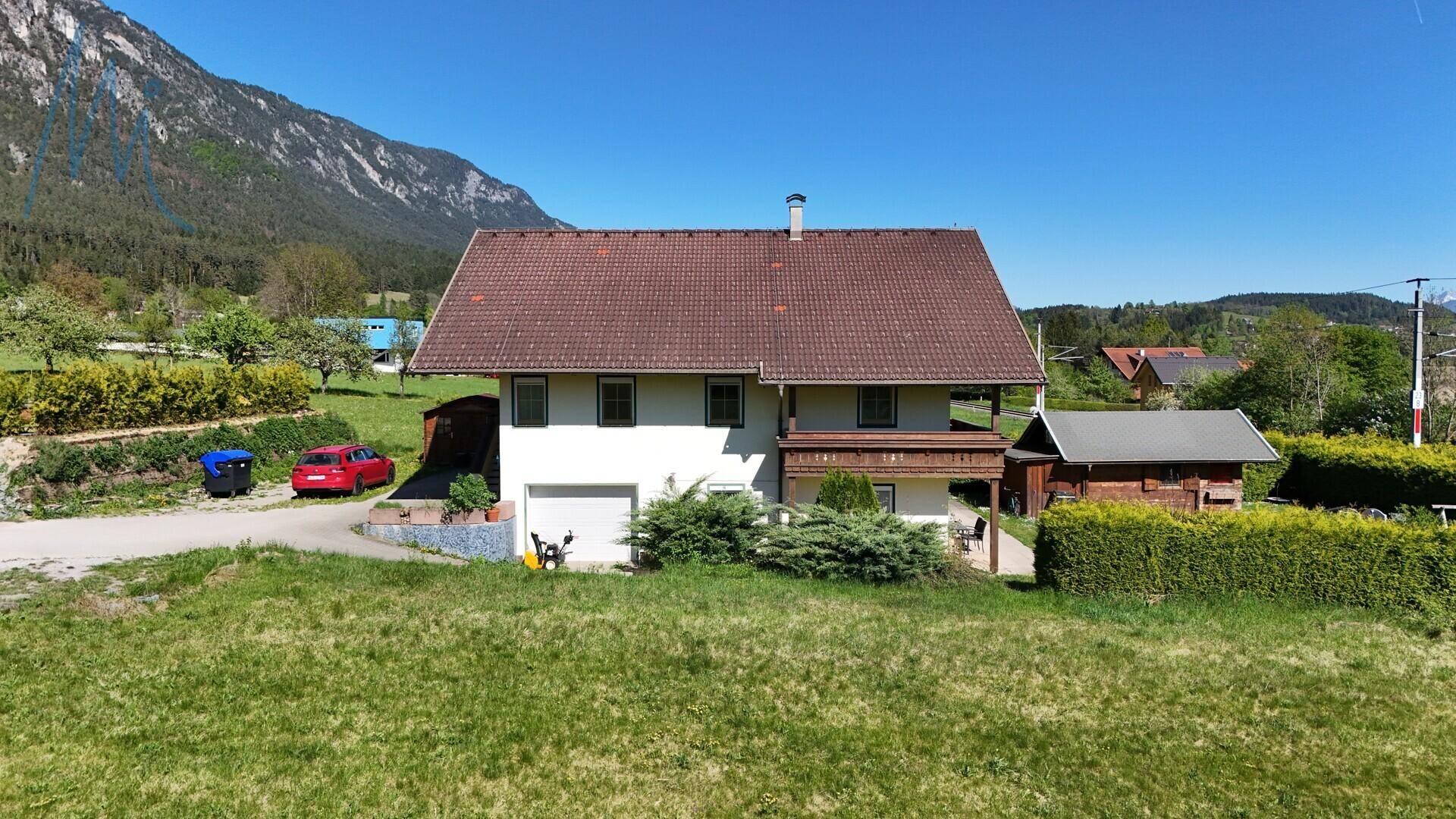 Hausfassade mit Garage und rotem Fahrzeug auf dem Grundstück, umgeben von grünen Wiesen und Bergen.