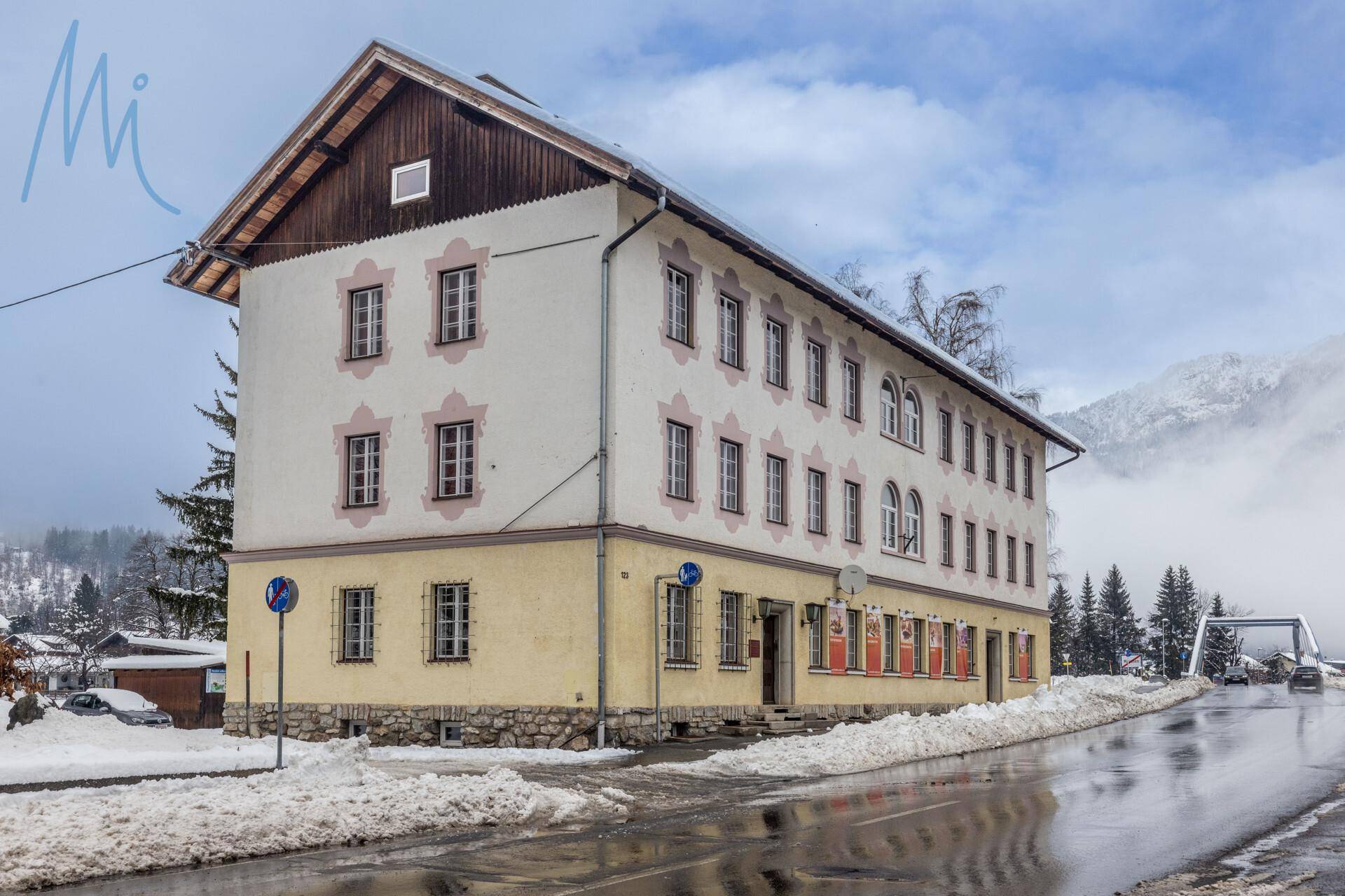 Traditionelles Gebäude mit mehreren Fenstern und schneebedeckter Straße im Vordergrund, umgeben von Bergen.