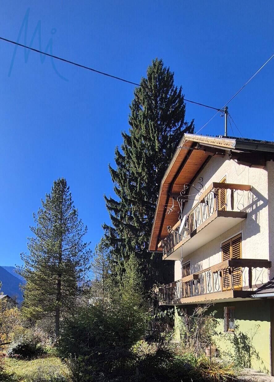 Traditionelles Haus mit Holzbalkonen und Fensterläden, umgeben von hohen Bäumen und Bergen unter blauem Himmel.