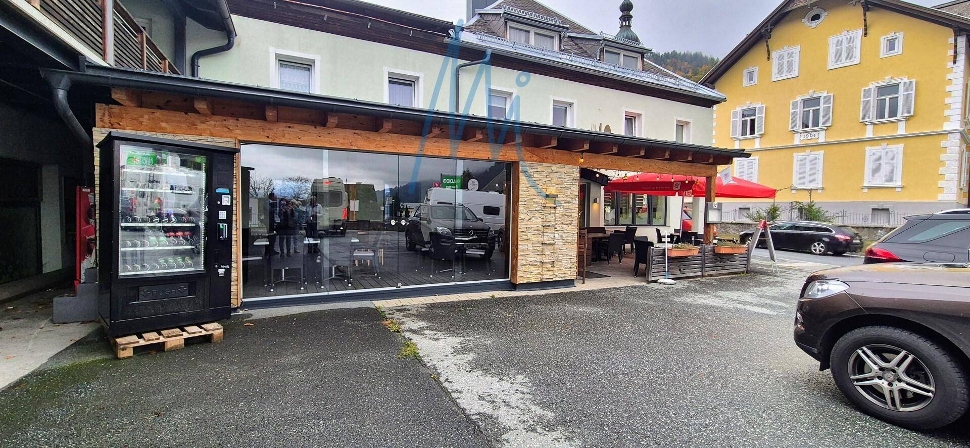 Außenansicht eines Gebäudes mit moderner Glasfassade, Vending-Automat und überdachtem Terrassenbereich mit Sitzgelegenheiten.