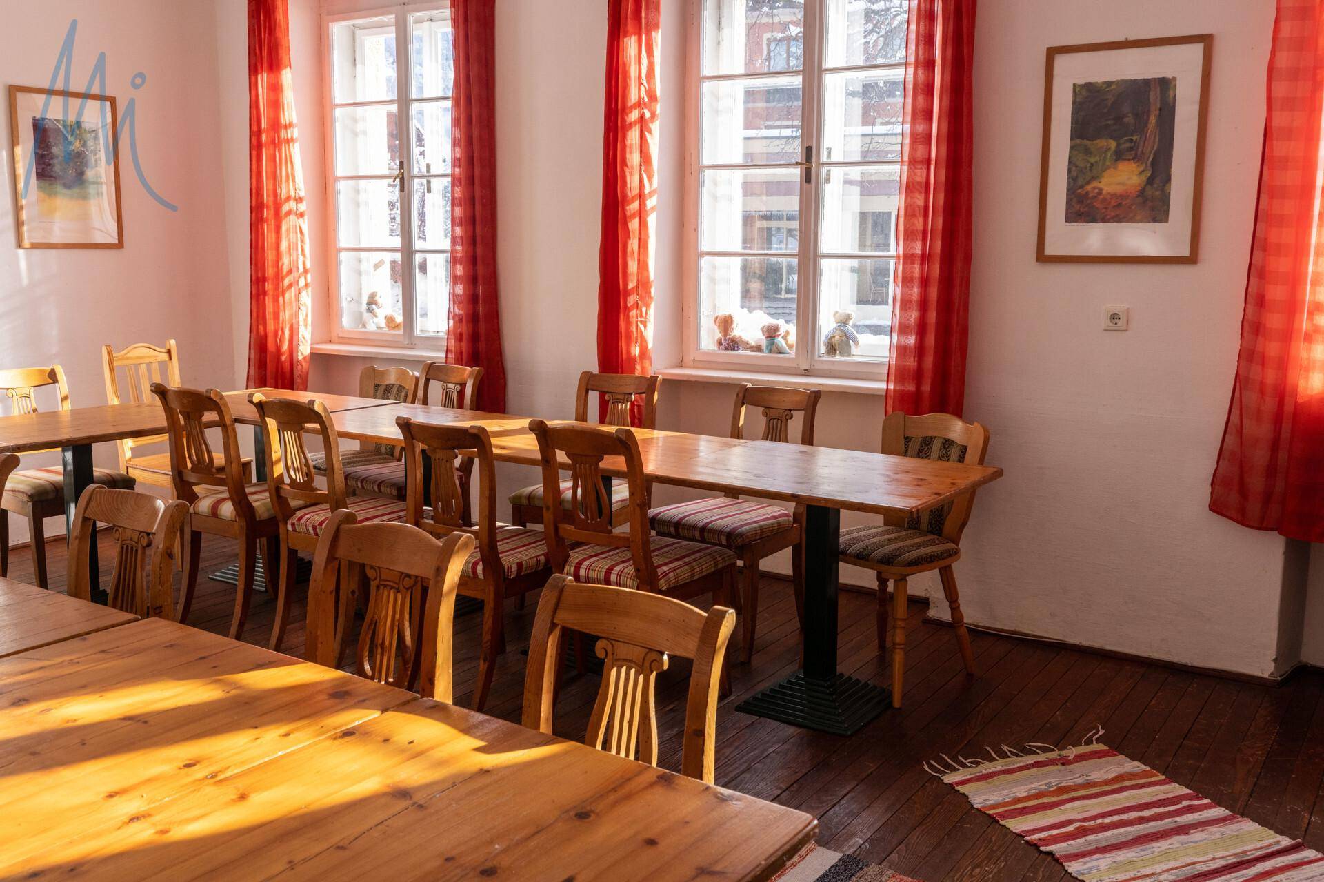 Heller Speisesaal mit Holztischen und Stühlen, roten Vorhängen und natürlichem Lichteinfall durch große Fenster.