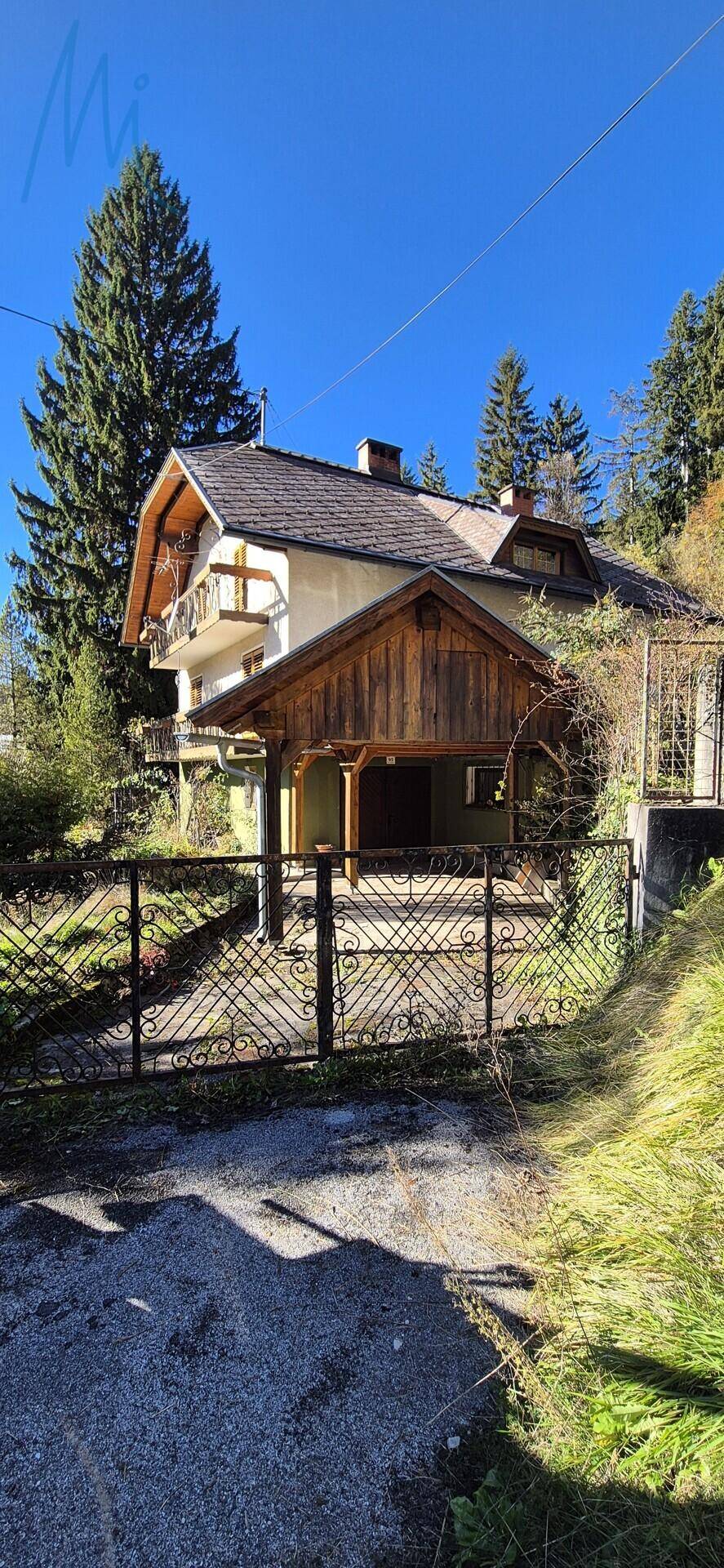 Haus mit Carport und dekorativem Metallzaun, umgeben von üppigem Grün, mit einem weiteren Gebäude im Hintergrund.