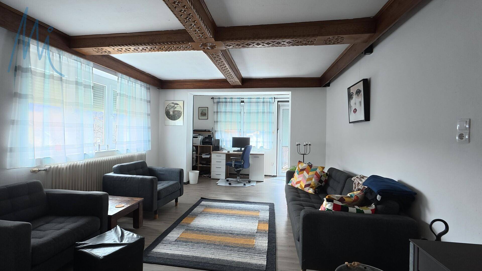 Geräumiges Wohnzimmer mit Holzbalkendecke, Sitzgelegenheiten und Zugang zum Arbeitszimmer.