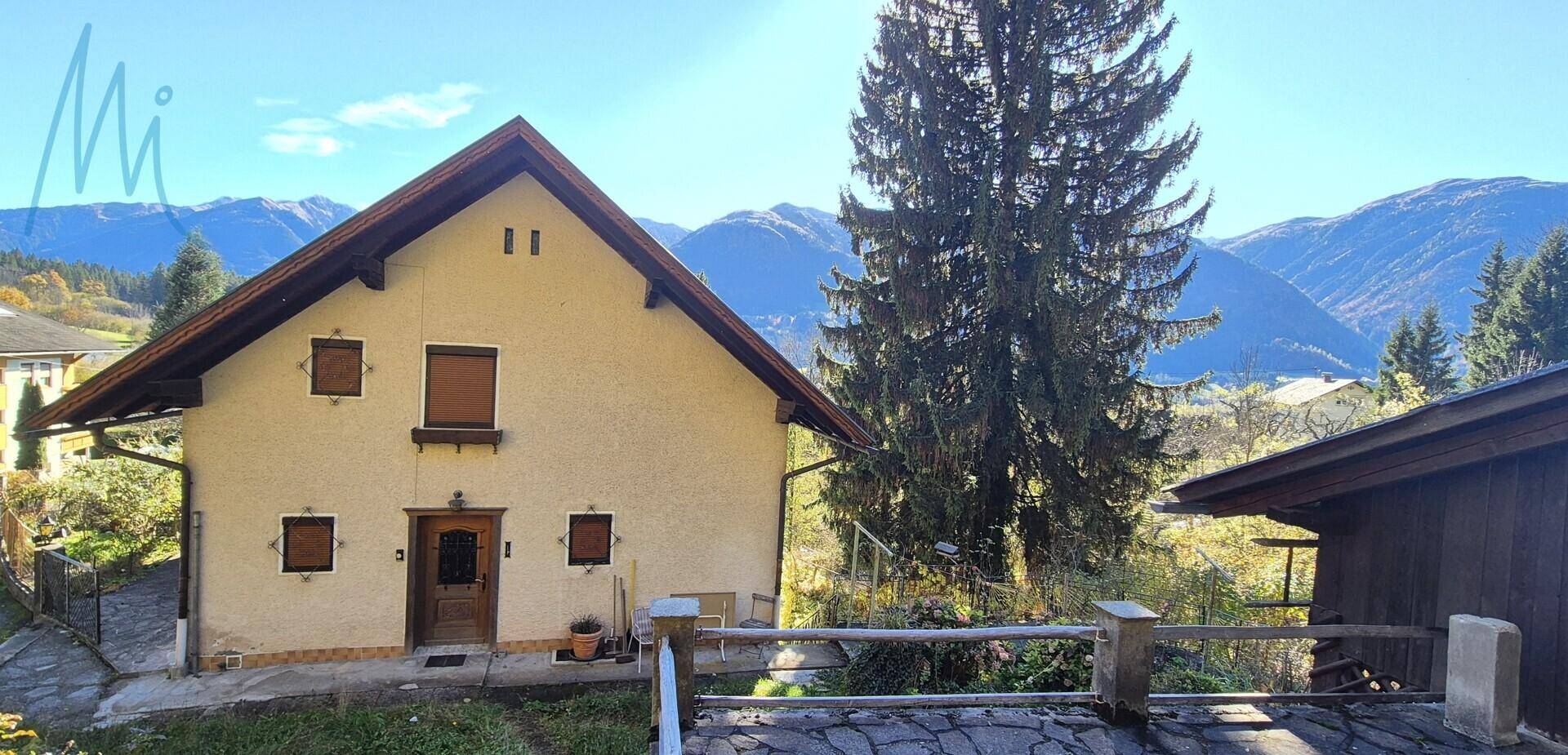 Haus mit heller Fassade und Satteldach, umgeben von Bäumen und einer beeindruckenden Bergkulisse.