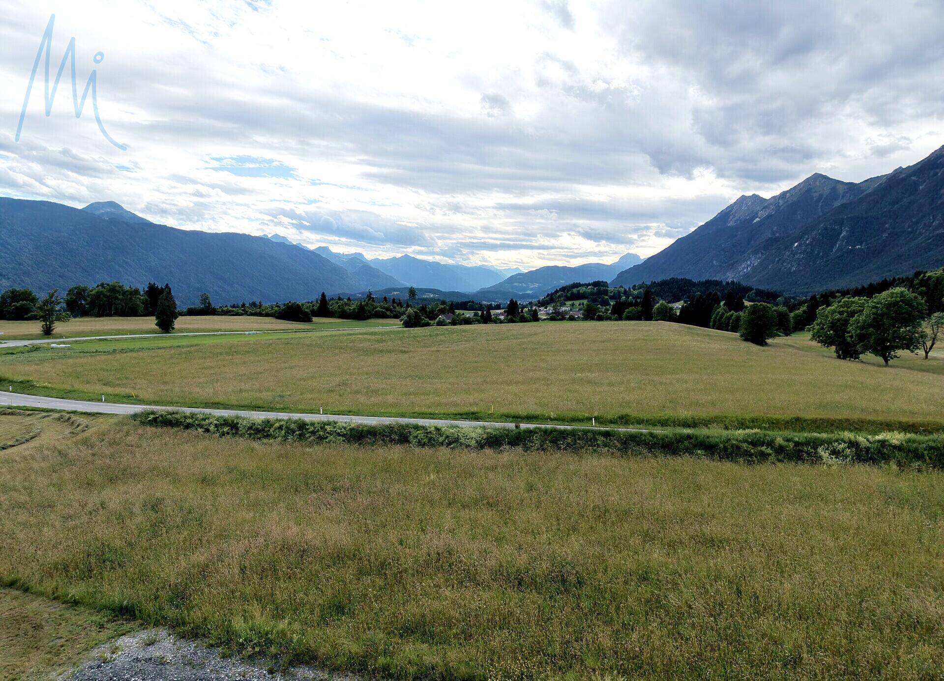 Weitläufige grüne Felder mit einer Straße im Vordergrund und majestätischen Bergen im Hintergrund.