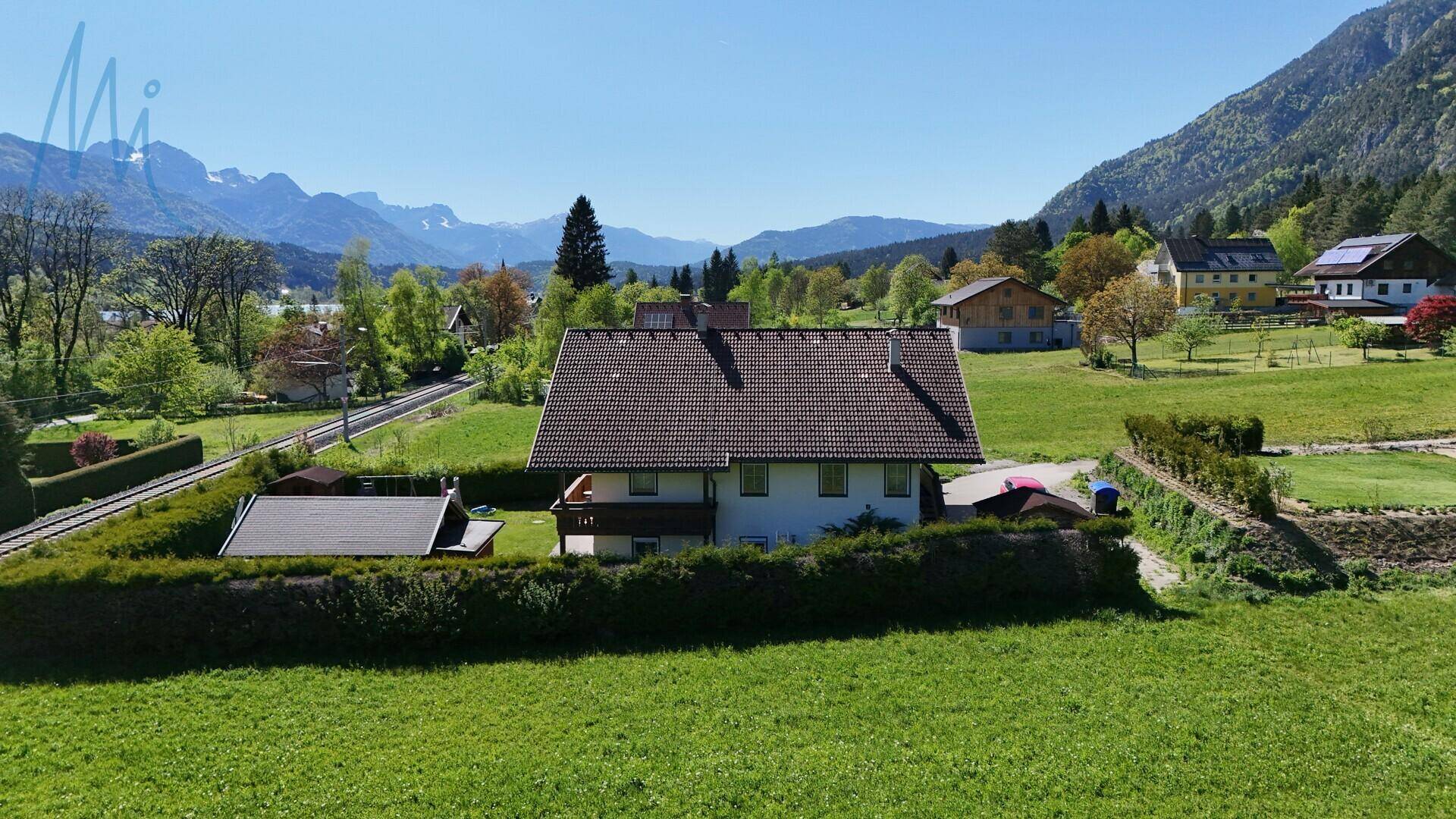 Luftaufnahme des Hauses in einer ländlichen Siedlung mit Bahnlinie und beeindruckendem Alpenpanorama.