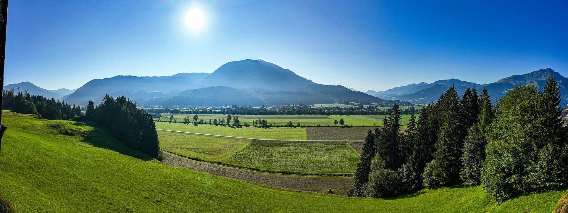 Panoramablick über weite grüne Felder und Wälder mit majestätischen Bergen im Hintergrund.