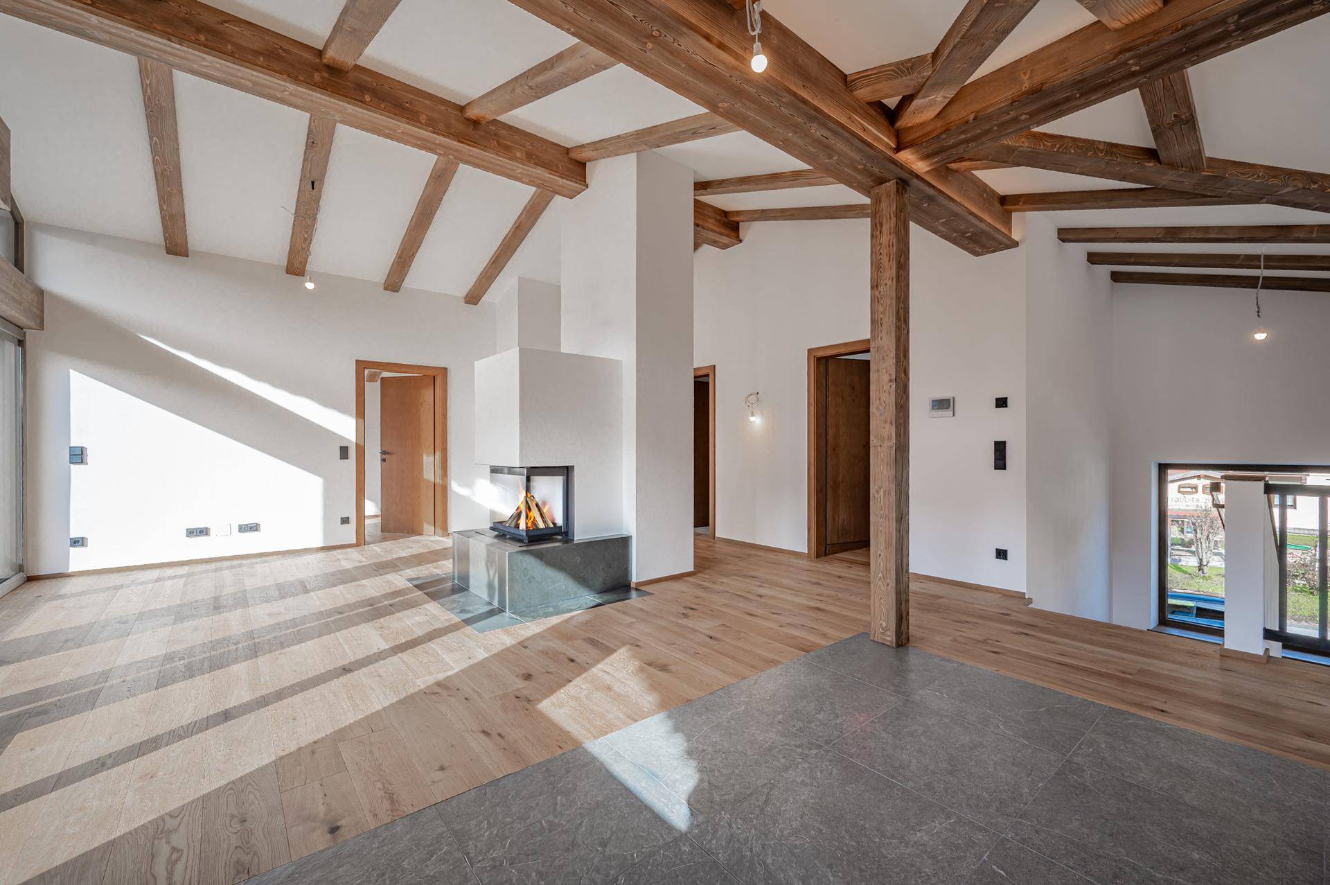 Heller Wohnbereich mit Holzbalken, modernem Kamin und unterschiedlichen Bodenbelägen.