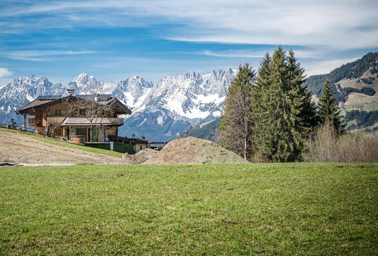 Alpines Chalet vor beeindruckender Kulisse schneebedeckter Berge und weiten Wiesen unter blauem Himmel.