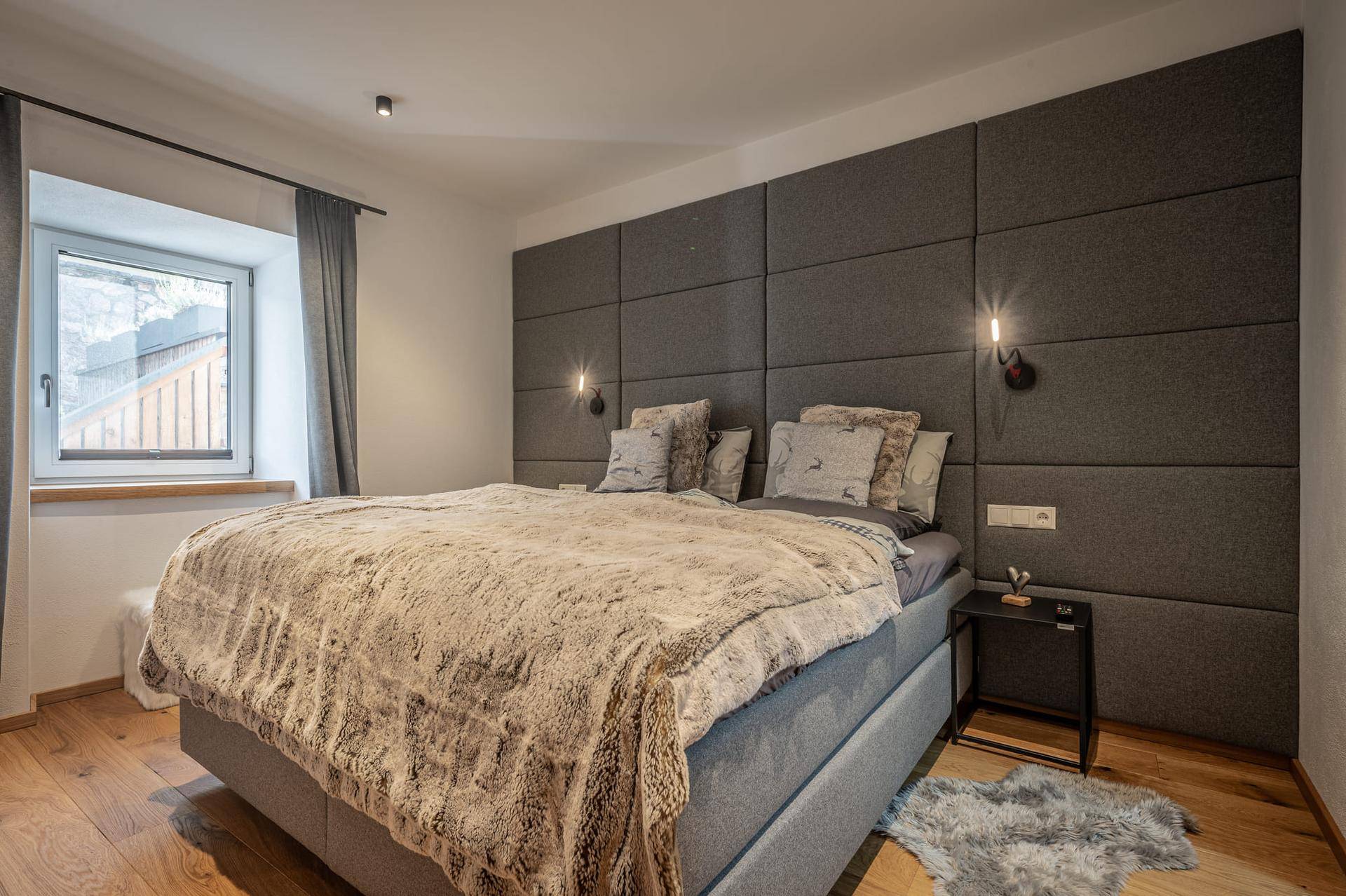 Modernes Schlafzimmer mit gepolsterter Wandverkleidung, Holzboden und natürlichem Lichteinfall durch das Fenster.