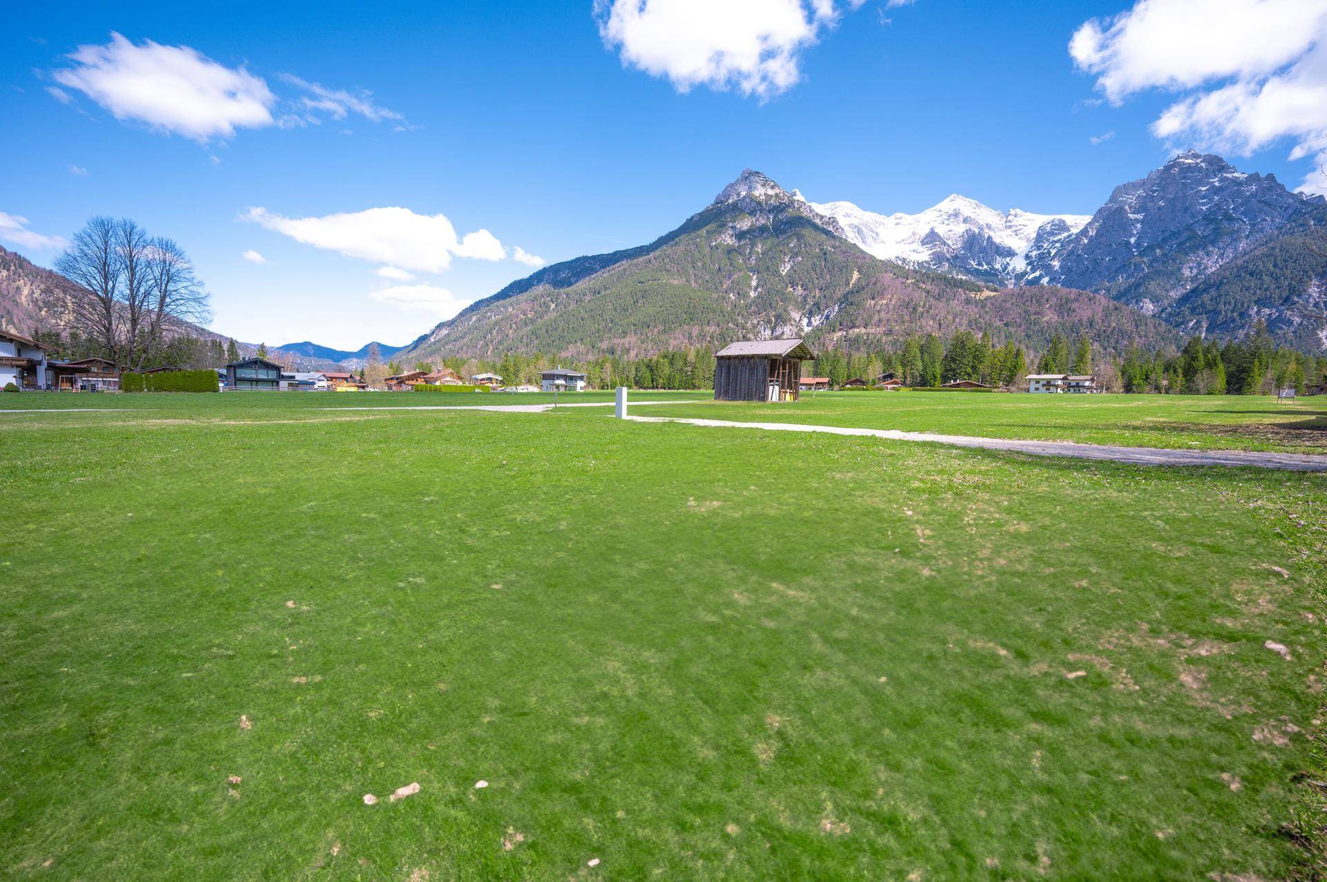 Panoramablick über eine weite grüne Wiese mit schneebedeckten Berggipfeln unter blauem Himmel.
