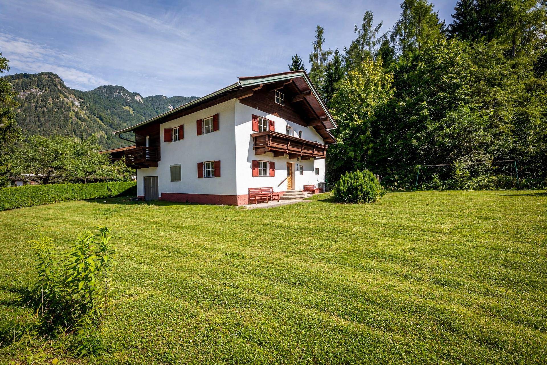 Charmantes Landhaus mit weitläufigem Rasen, umgeben von Bäumen und einer malerischen Berglandschaft.