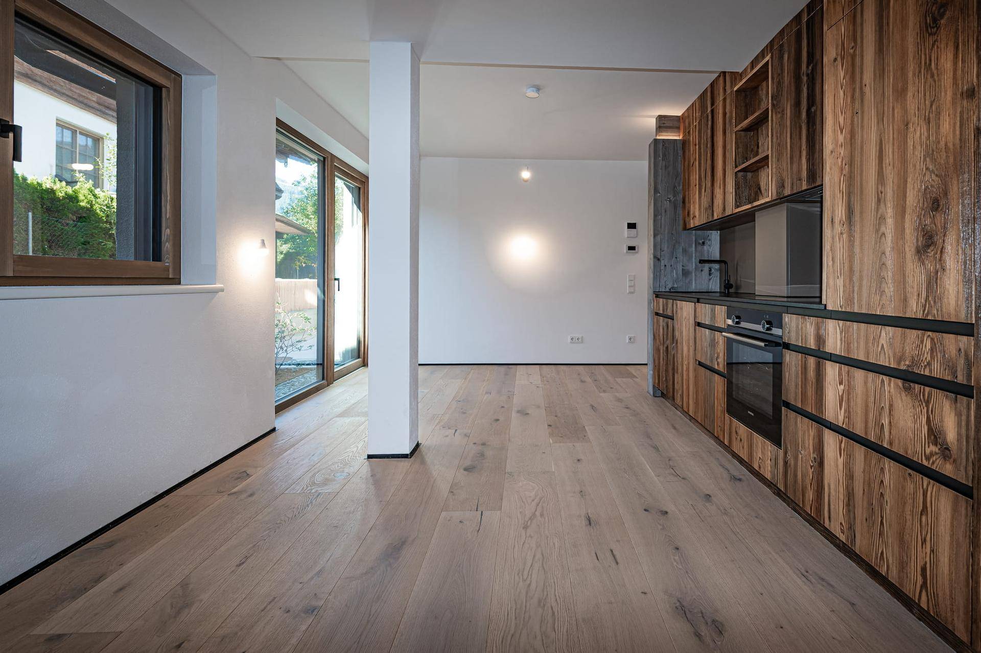 Heller Wohnbereich mit Holzboden und großen Fenstern, angrenzend an eine moderne Holzküche.