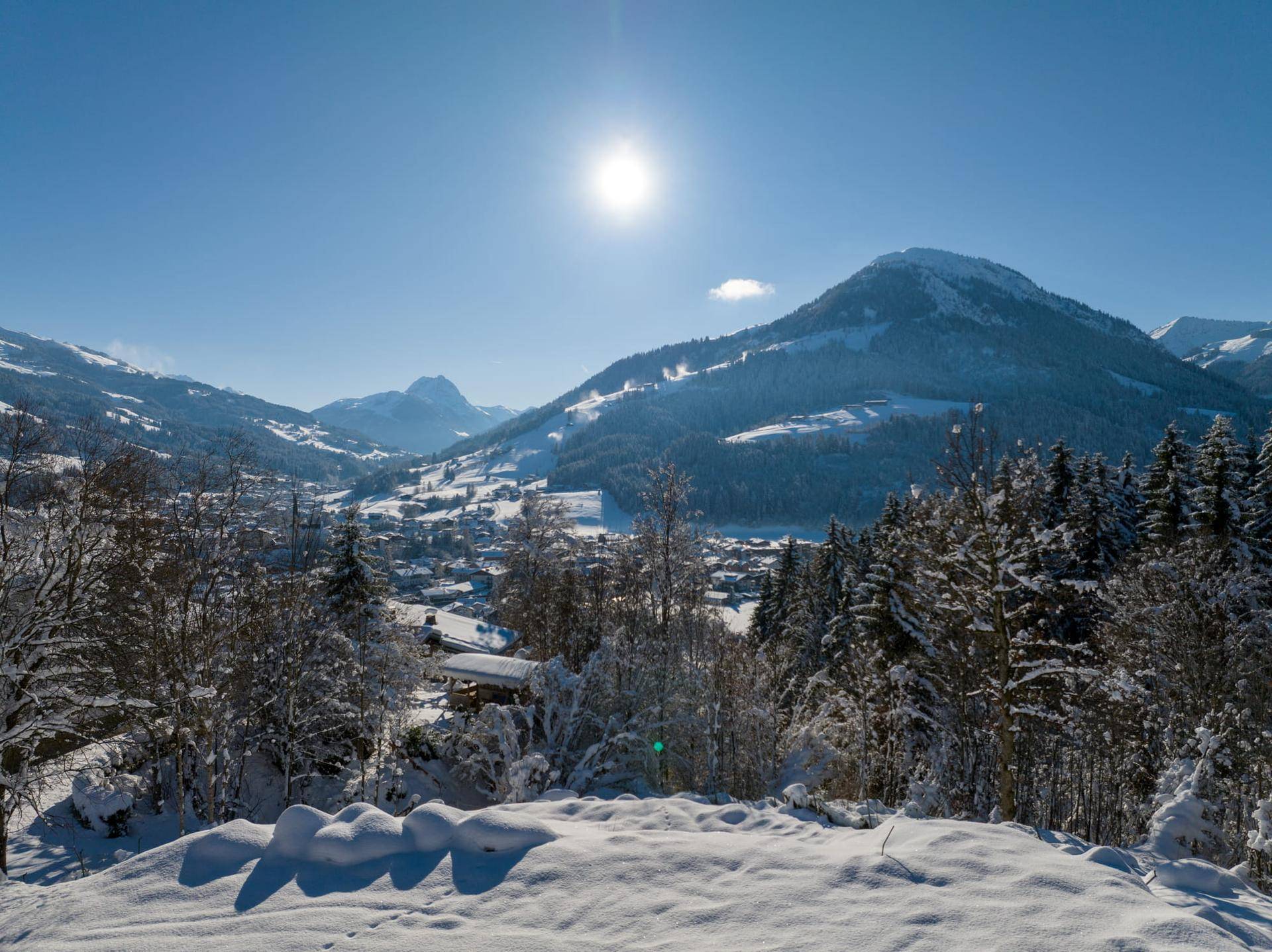 Weitläufige Winterlandschaft mit verschneitem Tal, Häusern und Bergen unter strahlendem Sonnenschein.