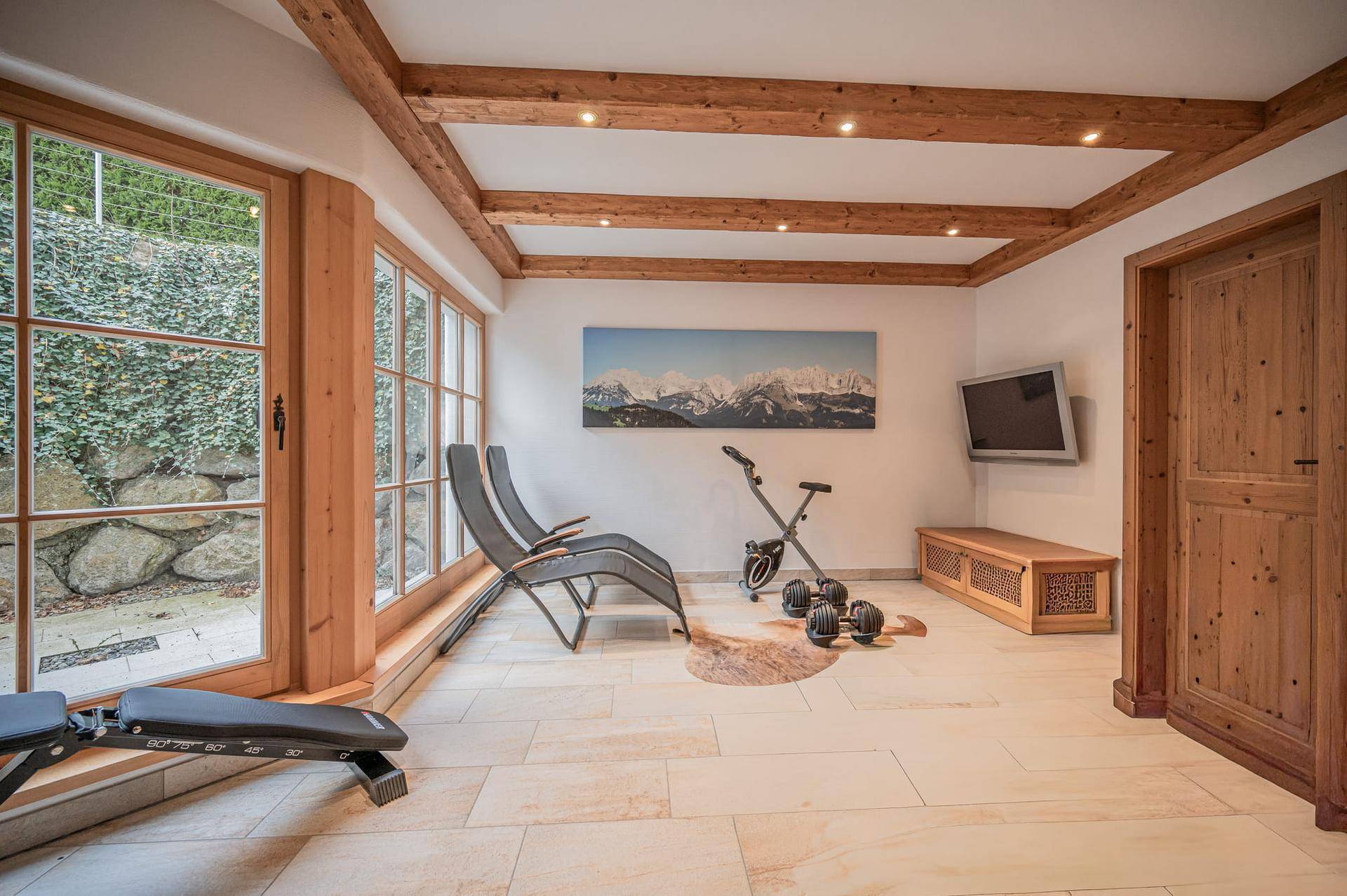 Fitnessraum mit Trainingsgeräten, Holzbalkendecke und großen Fenstern zum Garten.